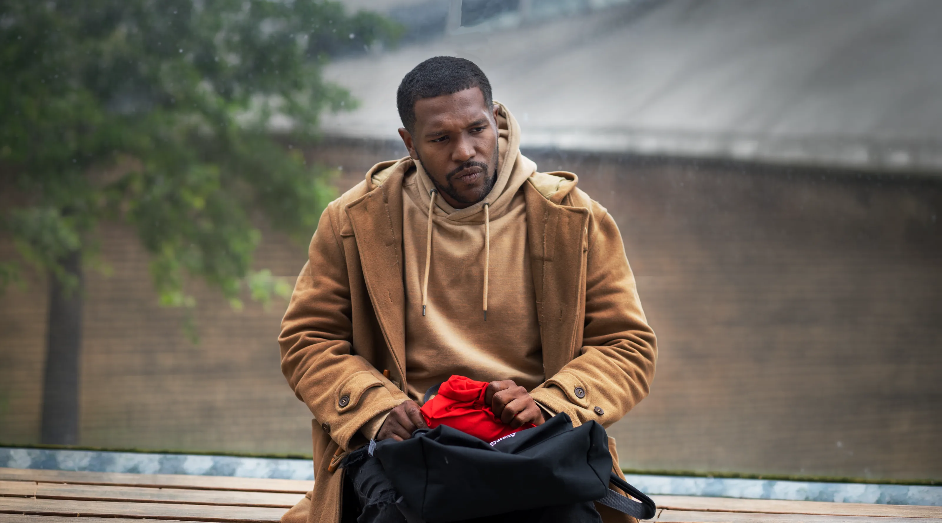Re:Tension: a man in a camel coat and hoodie sits on a bench, a red bag in his hands